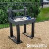 A wooden dog leash and collar station at a dog park, with leashes and collars placed on top. The station is next to a metal fence, with trees and wood chips on the ground. Text on the photo promotes Gyms for Dogs.