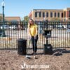A woman in a yellow shirt uses The Pet Waste Station - In-Ground Mount by a fence, cleaning up pet waste with a scooper. A black trash bin, bag dispenser, and "PET WASTE STATION" are on the ground in front of her outdoors.