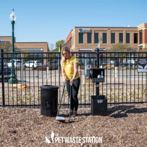 A woman in a yellow shirt uses The Ultimate Park Caddy System—including Waste Caddy & Storage Caddy with Scooper—to clean pet waste at a dog park by a black waste station and trash bin, with buildings and a fence in the background.