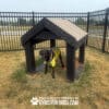 A black dog in a yellow shirt stands inside a Tunnel House (L/X) on brown grass at a dog park, surrounded by a black metal fence. Text at the bottom reads, "PLAY & AGILITY POWERED BY GYMS FOR DOGS.COM.
