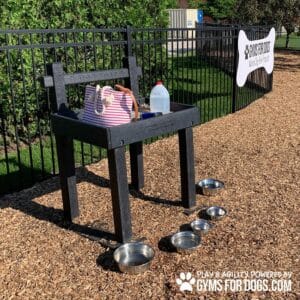 A black Dog Post - Leash Holder & Toy Box with metal bowls, striped bag, and water jug sits on wood chips near a black fence, with a "Gyms for Dogs" Dog Park sign visible in the background.