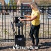 A woman in a yellow shirt dispenses a dog waste bag from a black pet waste station near a fenced area in an outdoor park. The station has instructions and a sign reading "NO WASTE" with a dog icon.