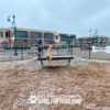 Two dogs play on Ellie's Boulder Bridge Pro—Bridge, Set of 2 Rocks, and Bumper Pads—in a fenced dog park. One stands on the bridge while the other leaps off onto mulch, buildings visible in the background on an overcast day.