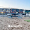 Several dogs and people enjoy an outdoor dog park with businesses in the background. The park features Ellie's Boulder Bridge Pro—Bridge, Set of 2 Rocks, and Bumper Pads—and a “Gyms for Dogs” sign.