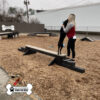 A woman stands on a Jump Balance Beam in a dog park, hugging a black dog with its front paws on the beam. Wood chips cover the ground, benches and another dog are visible in the background. The Gyms For Dogs logo appears in the corner.