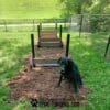 A black dog sits on a mulched path in front of a Jump Hurdle Run - Set of 3 agility obstacle at a fenced Dog Park, with "GYMSFORDOGS.COM" text displayed at the bottom of the image.