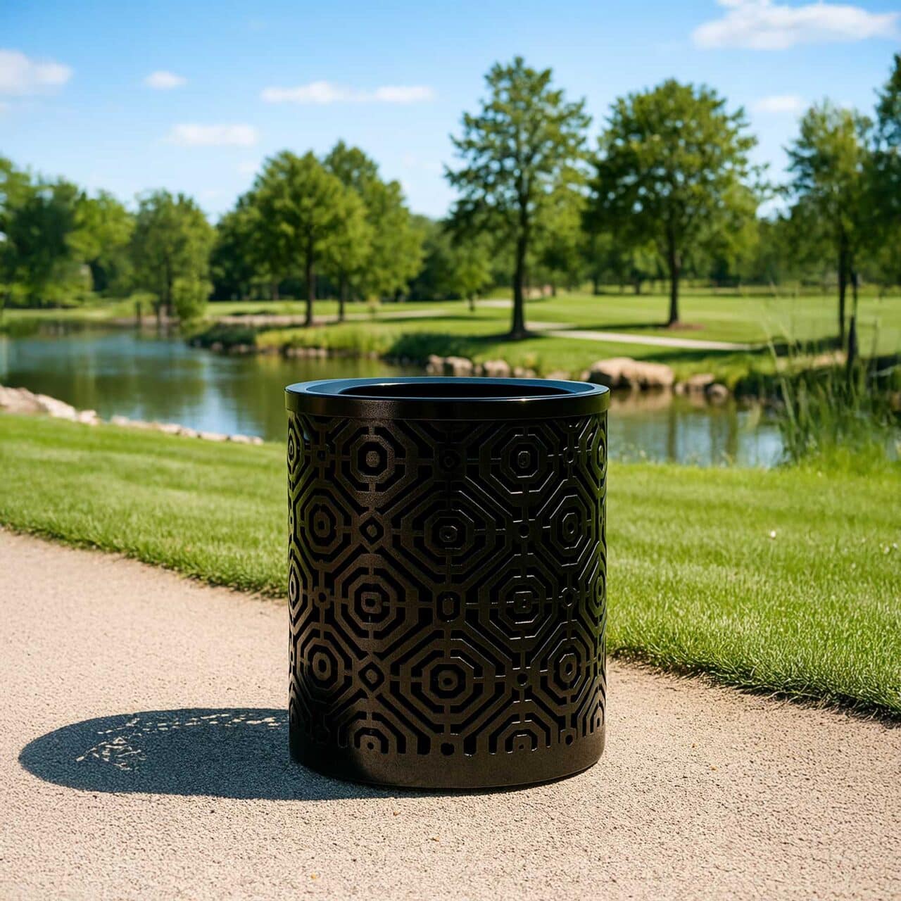 A black trash can with a flat-top lid, sunlight casting shadows on a pathway by a small pond in a green park.