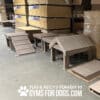 A warehouse with stacks of cardboard boxes contains several brown wooden dog agility ramps and platforms for a Dog Park. The text reads: "PLAY & AGILITY POWERED BY GYMSFORDOGS.COM" with a paw print.