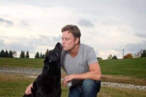A man crouches on grass, smiling as a black dog stands on its hind legs and licks his face. They are outdoors in a dog park with green grass, trees, and a cloudy sky in the background.